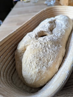A loaf of unbaked bread dough dusted with flour rests in a wooden banneton, with a close-up view highlighting its texture and shape. The background is a blurred wooden table with a few scattered items.