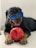A Rottweiler wearing blue-tinted sunglasses lies on the floor, playfully holding a large red toy in its front paws. The dog's tongue is out, suggesting a relaxed and happy demeanor.