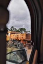A large dump truck carrying a load of rocks is visible through the window of another vehicle. The truck is on a dusty road, causing some dust to billow around its tires. The surrounding area includes green foliage and an overcast sky.