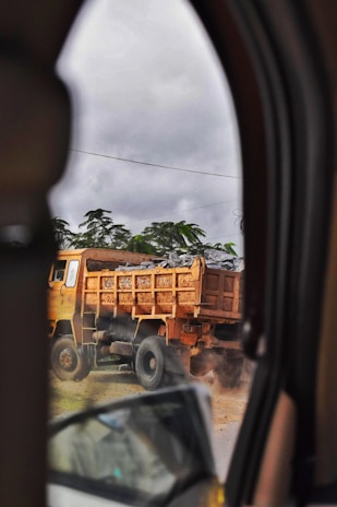 A large dump truck carrying a load of rocks is visible through the window of another vehicle. The truck is on a dusty road, causing some dust to billow around its tires. The surrounding area includes green foliage and an overcast sky.