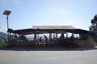 A small roadside structure with a corrugated metal roof, housing several people engaged in various activities. It is situated in a rural landscape with hills and greenery in the background. The structure appears to be made of wood and bricks. There is a solar panel mounted on a pole nearby.