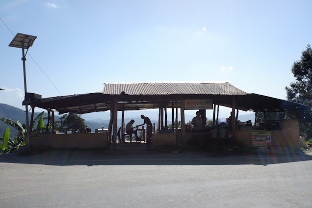 A small roadside structure with a corrugated metal roof, housing several people engaged in various activities. It is situated in a rural landscape with hills and greenery in the background. The structure appears to be made of wood and bricks. There is a solar panel mounted on a pole nearby.