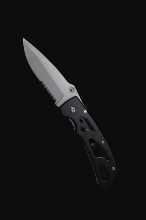 A folding knife with a partially serrated blade and a black handle is set against a stark black background. The handle has cutout designs that contribute to its lightweight appearance.