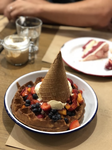 A waffle topped with a scoop of ice cream, a waffle cone, and an assortment of fresh fruits like strawberries, blueberries, and possibly peaches, served on a white plate with a blue rim. In the background, there is a blurred image of another dessert on a separate plate, along with two glass cups containing a beverage, possibly coffee or similar drink. A person's arm can also be seen resting on the table.