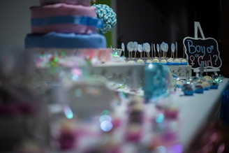 A decorative table is set with a variety of colorful treats, including a tiered cake with pink and blue layers. There are cake pops with designs, pastel cupcakes, and a small chalkboard sign that reads 'Boy or Girl?'. Sparkling decorations adorn the table, suggesting a festive atmosphere.