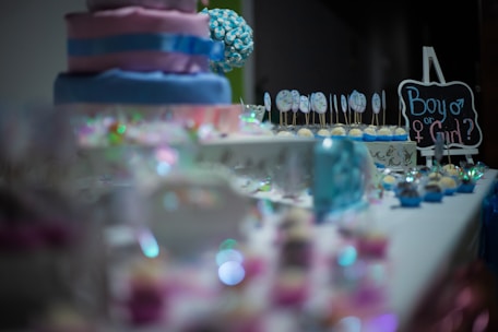 A decorative table is set with a variety of colorful treats, including a tiered cake with pink and blue layers. There are cake pops with designs, pastel cupcakes, and a small chalkboard sign that reads 'Boy or Girl?'. Sparkling decorations adorn the table, suggesting a festive atmosphere.