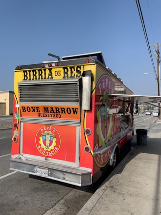A vibrant food truck parked on the side of the road, featuring a bright red color with yellow accents and signs advertising 'Birria de Res' and 'Bone Marrow Tacos.' The truck has a prominent logo with a bull's skull and the name 'Pepe's Red Tacos.' An open serving window is visible on the side.