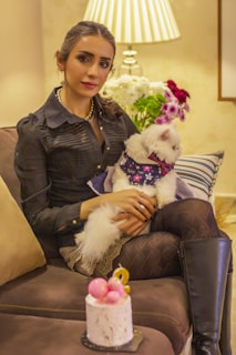 A woman with dark hair and a black blouse is sitting on a sofa, holding a white fluffy dog dressed in a patterned outfit. Next to her is a round table with a small cake topped with decorative pink and yellow elements. Behind them, there is a lamp and a bouquet of white, pink, and green flowers adding a cozy atmosphere.