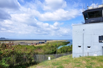 A control tower is situated on the right side overlooking an airport runway. An aircraft is parked on the runway surrounded by various airport maintenance vehicles. The scene includes lush greenery, a fence in the foreground, and a river with a bend leading towards the open ocean under a partly cloudy sky.