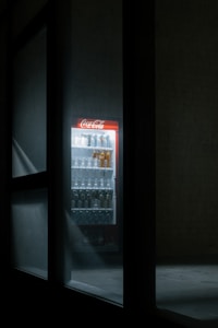 A dimly lit vending machine with a bright Coca-Cola sign is visible through a window. The machine is stocked with various drinks in bottles and cans, and the surrounding area is in shadow, adding a mysterious atmosphere.