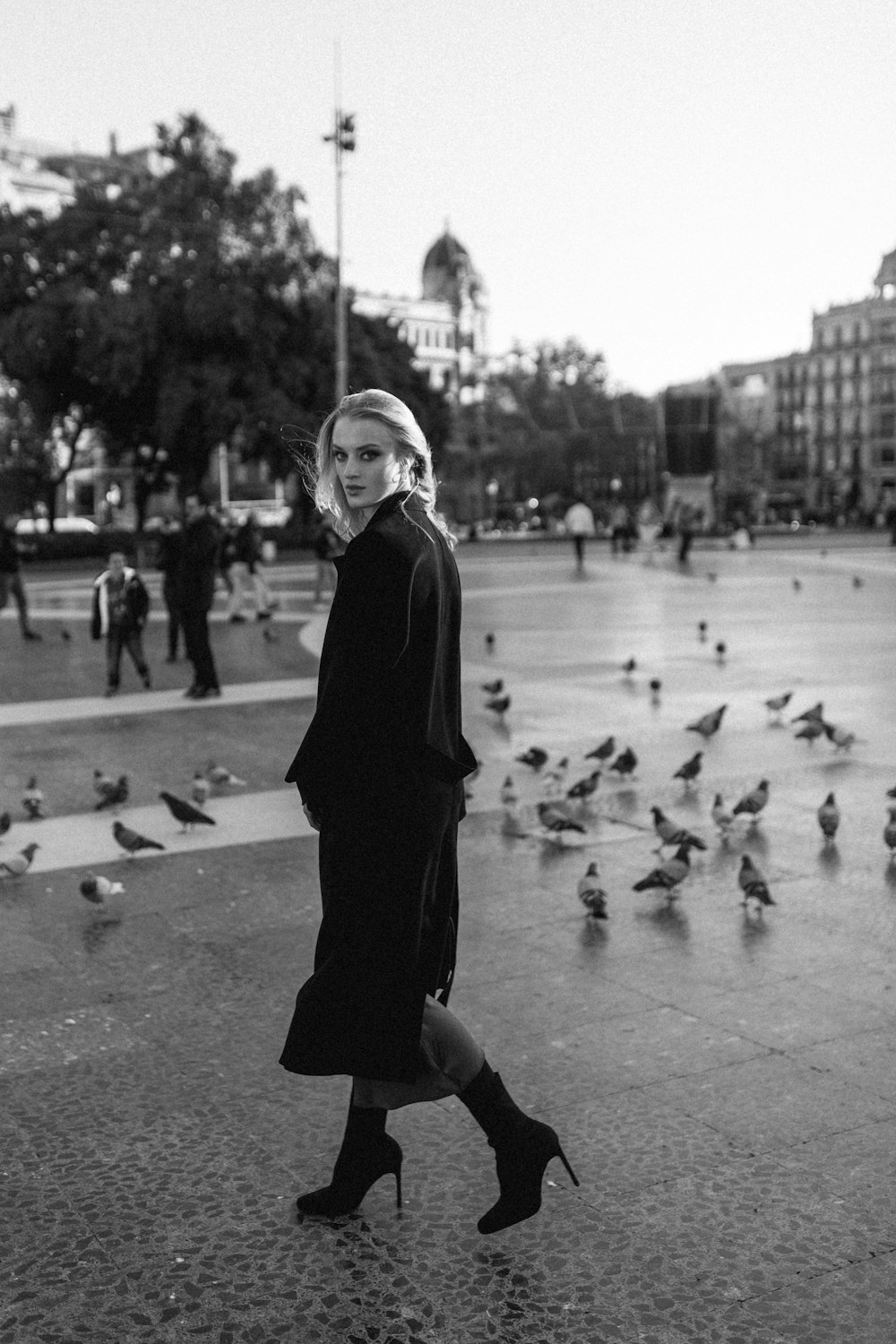 a woman in a black coat is standing in front of a flock of birds