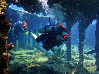 Two scuba divers explore an underwater structure, likely a shipwreck or a coral-covered framework. Sunlight filters through the water, casting a serene glow on the scene. The divers are equipped with scuba gear including tanks, masks, and fins.