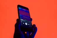 A hand holds a smartphone displaying a music player app with a song titled 'Change' by 'Like Crashing Waves'. The background is a vibrant orange, creating a striking contrast with the phone's dark silhouette and screen.