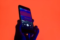 A hand holds a smartphone displaying a music player app with a song titled 'Change' by 'Like Crashing Waves'. The background is a vibrant orange, creating a striking contrast with the phone's dark silhouette and screen.