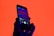 A hand holds a smartphone displaying a music player app with a song titled 'Change' by 'Like Crashing Waves'. The background is a vibrant orange, creating a striking contrast with the phone's dark silhouette and screen.