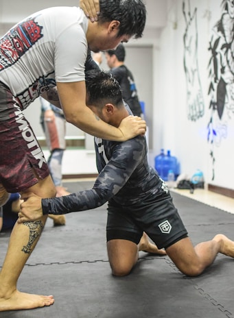 Two individuals are engaged in a grappling match on a mat. The person on the left is standing upright with their arm around the neck of the kneeling individual, who appears to be executing a wrestling move.