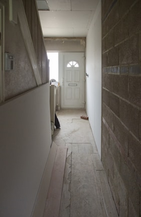 A narrow hallway with unfinished walls. The floor appears to be wooden and partially covered with construction materials. At the end of the hallway is a closed white door with a semi-circular window at the top. The right wall is made of exposed brick, and there are various construction items scattered around.