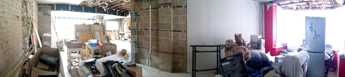 A partially renovated interior space with exposed bricks and beams. The area is cluttered with construction materials, cardboard boxes, and various tools. On the right, there is a small sitting area with a refrigerator, a stuffed animal, and other household items.