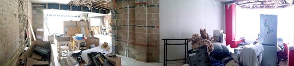 A partially renovated interior space with exposed bricks and beams. The area is cluttered with construction materials, cardboard boxes, and various tools. On the right, there is a small sitting area with a refrigerator, a stuffed animal, and other household items.