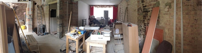 A large room in the midst of renovation, filled with various tools, construction materials, and furniture pieces. Exposed brick walls are visible, and there is a combination of unfinished and partially completed sections of the interior. Sunlight filters in through a window at the far end, where red curtains are hung.