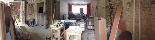 A large room in the midst of renovation, filled with various tools, construction materials, and furniture pieces. Exposed brick walls are visible, and there is a combination of unfinished and partially completed sections of the interior. Sunlight filters in through a window at the far end, where red curtains are hung.