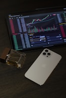 An iPad displays a cryptocurrency trading platform with various charts and numbers. Next to the iPad, there is a white smartphone laid flat on a dark, textured surface. A square glass perfume bottle with some liquid inside is positioned beside the smartphone.