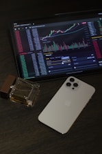 An iPad displays a cryptocurrency trading platform with various charts and numbers. Next to the iPad, there is a white smartphone laid flat on a dark, textured surface. A square glass perfume bottle with some liquid inside is positioned beside the smartphone.