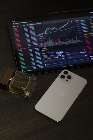 An iPad displays a cryptocurrency trading platform with various charts and numbers. Next to the iPad, there is a white smartphone laid flat on a dark, textured surface. A square glass perfume bottle with some liquid inside is positioned beside the smartphone.