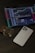 An iPad displays a cryptocurrency trading platform with various charts and numbers. Next to the iPad, there is a white smartphone laid flat on a dark, textured surface. A square glass perfume bottle with some liquid inside is positioned beside the smartphone.