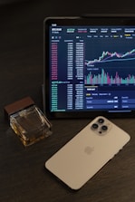 A tablet displays a cryptocurrency trading chart with various data points and graphs. Next to it, there is a smartphone with a metallic back and multiple camera lenses. A cologne bottle with a wooden cap is placed beside these devices on a dark wooden surface.