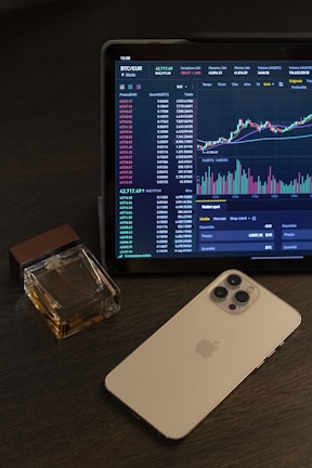 A tablet displays a cryptocurrency trading chart with various data points and graphs. Next to it, there is a smartphone with a metallic back and multiple camera lenses. A cologne bottle with a wooden cap is placed beside these devices on a dark wooden surface.