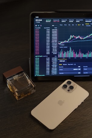 A tablet displays a cryptocurrency trading chart with various data points and graphs. Next to it, there is a smartphone with a metallic back and multiple camera lenses. A cologne bottle with a wooden cap is placed beside these devices on a dark wooden surface.
