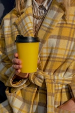 A person wearing a yellow and brown plaid coat holds a yellow coffee cup with a black lid. The coat is layered over a striped shirt and a gold chain necklace can be seen. The style appears warm and fashionable.