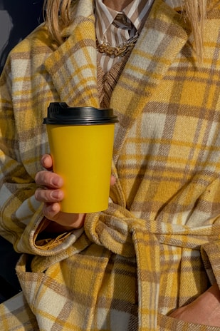 A person wearing a yellow and brown plaid coat holds a yellow coffee cup with a black lid. The coat is layered over a striped shirt and a gold chain necklace can be seen. The style appears warm and fashionable.