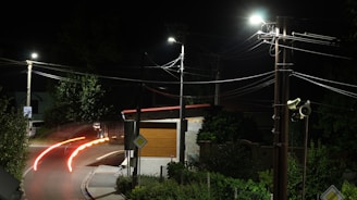 A nighttime scene featuring a suburban street corner illuminated by streetlights. Light trails from vehicles create streaks along the road, indicating movement. Power lines crisscross the scene above, and there are trees and a small building with a brown door or shutter.