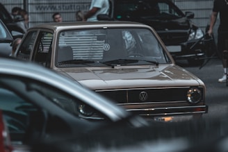 A vintage Volkswagen car is parked among several modern vehicles on a street. People are gathered around, some standing and others partially visible in the background. The atmosphere appears urban and slightly crowded.