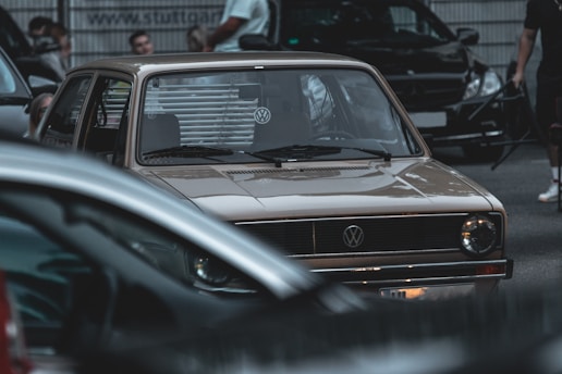 A vintage Volkswagen car is parked among several modern vehicles on a street. People are gathered around, some standing and others partially visible in the background. The atmosphere appears urban and slightly crowded.
