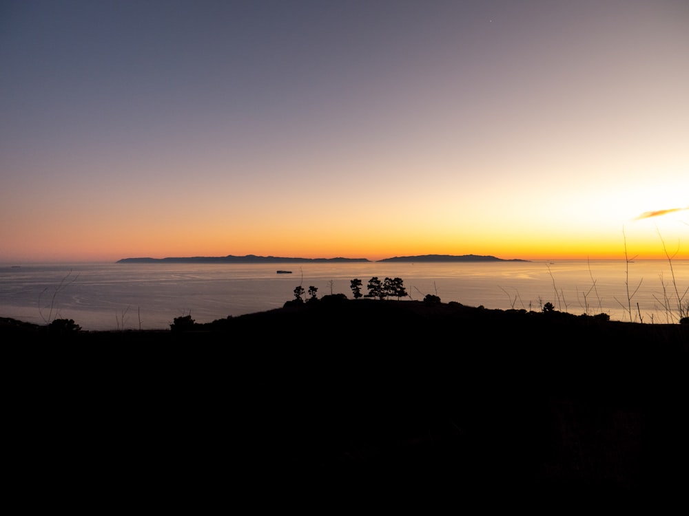 the sun is setting over the ocean with a hill in the foreground