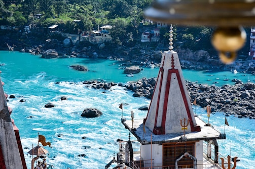 A serene riverscape featuring a vibrant turquoise river flowing over rocks. A temple with a conical spire and decorative flags stands prominently in the foreground, nestled among lush greenery and rustic buildings in the background.
