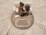 A small figurine display shows two figures sitting on a sofa on a wooden base with intricate lace around it. One figure places a comforting hand on the other’s arm, symbolizing support and togetherness. A plaque with an inspirational quote about encouragement and prayer is visible.