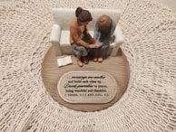 A small figurine display shows two figures sitting on a sofa on a wooden base with intricate lace around it. One figure places a comforting hand on the other’s arm, symbolizing support and togetherness. A plaque with an inspirational quote about encouragement and prayer is visible.