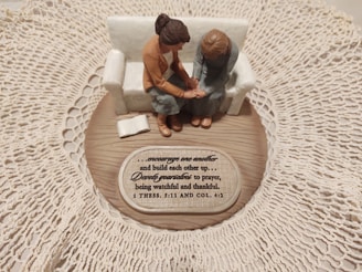 A small figurine display shows two figures sitting on a sofa on a wooden base with intricate lace around it. One figure places a comforting hand on the other’s arm, symbolizing support and togetherness. A plaque with an inspirational quote about encouragement and prayer is visible.