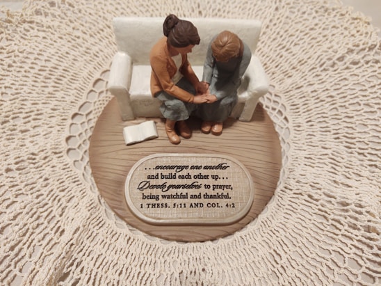 A small figurine display shows two figures sitting on a sofa on a wooden base with intricate lace around it. One figure places a comforting hand on the other’s arm, symbolizing support and togetherness. A plaque with an inspirational quote about encouragement and prayer is visible.