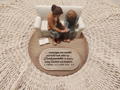 A small figurine display shows two figures sitting on a sofa on a wooden base with intricate lace around it. One figure places a comforting hand on the other’s arm, symbolizing support and togetherness. A plaque with an inspirational quote about encouragement and prayer is visible.