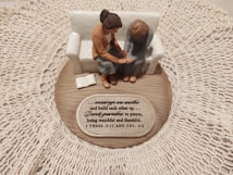 A small figurine display shows two figures sitting on a sofa on a wooden base with intricate lace around it. One figure places a comforting hand on the other’s arm, symbolizing support and togetherness. A plaque with an inspirational quote about encouragement and prayer is visible.