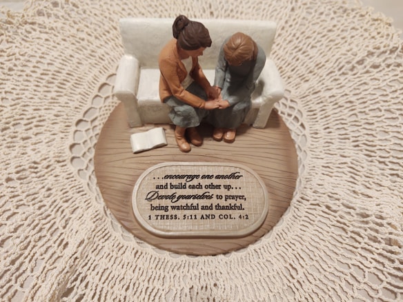 A small figurine display shows two figures sitting on a sofa on a wooden base with intricate lace around it. One figure places a comforting hand on the other&rsquo;s arm, symbolizing support and togetherness. A plaque with an inspirational quote about encouragement and prayer is visible.