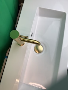 A modern and sleek bathroom sink with a shiny gold faucet. The sink is white and rectangular in shape, with a smooth and minimalist design. The surrounding area appears to be clean and well-lit, with a green backdrop visible on one side.