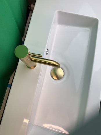 A modern and sleek bathroom sink with a shiny gold faucet. The sink is white and rectangular in shape, with a smooth and minimalist design. The surrounding area appears to be clean and well-lit, with a green backdrop visible on one side.