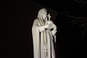 A white statue of a robed figure holding a globe topped with a cross. The figure has a calm expression and is draped in a rosary. The statue is illuminated against a dark background with faint outlines of tree branches.