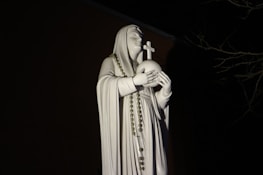 A white statue of a robed figure holding a globe topped with a cross. The figure has a calm expression and is draped in a rosary. The statue is illuminated against a dark background with faint outlines of tree branches.